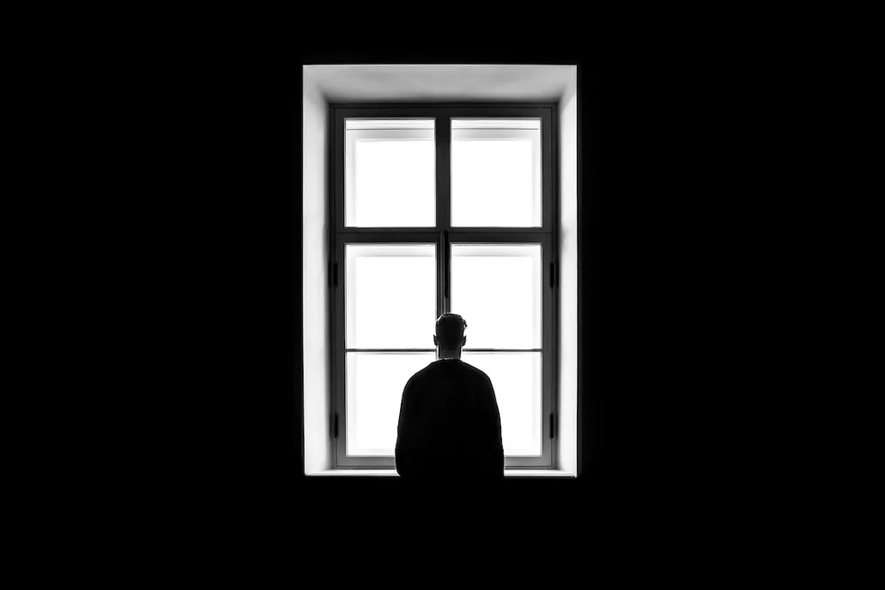 The image is completely dark except for a bright window and a man looking through it. We view him from behind. He's covered in shadow.
