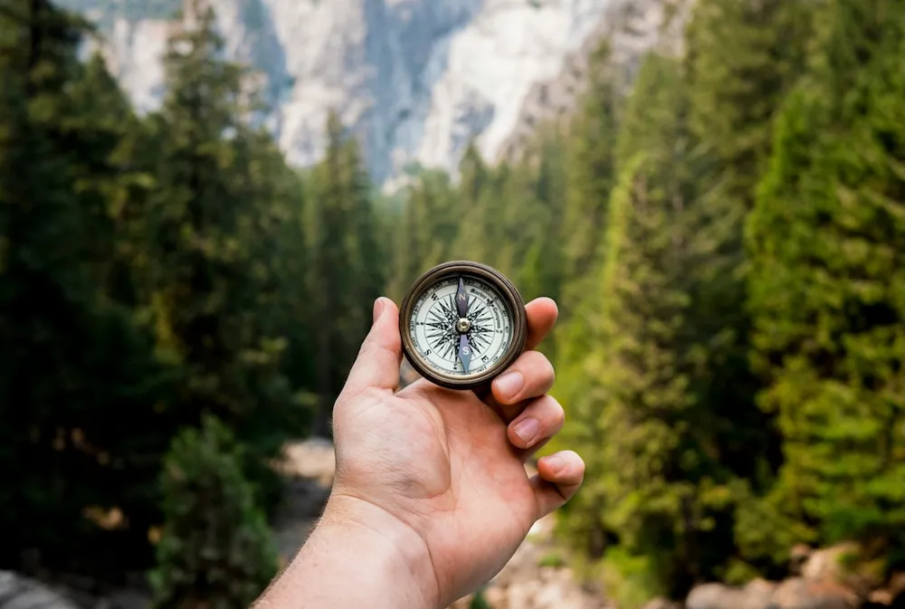 A compass shown in a man's hand with mountainous terrain as a backdrop.
